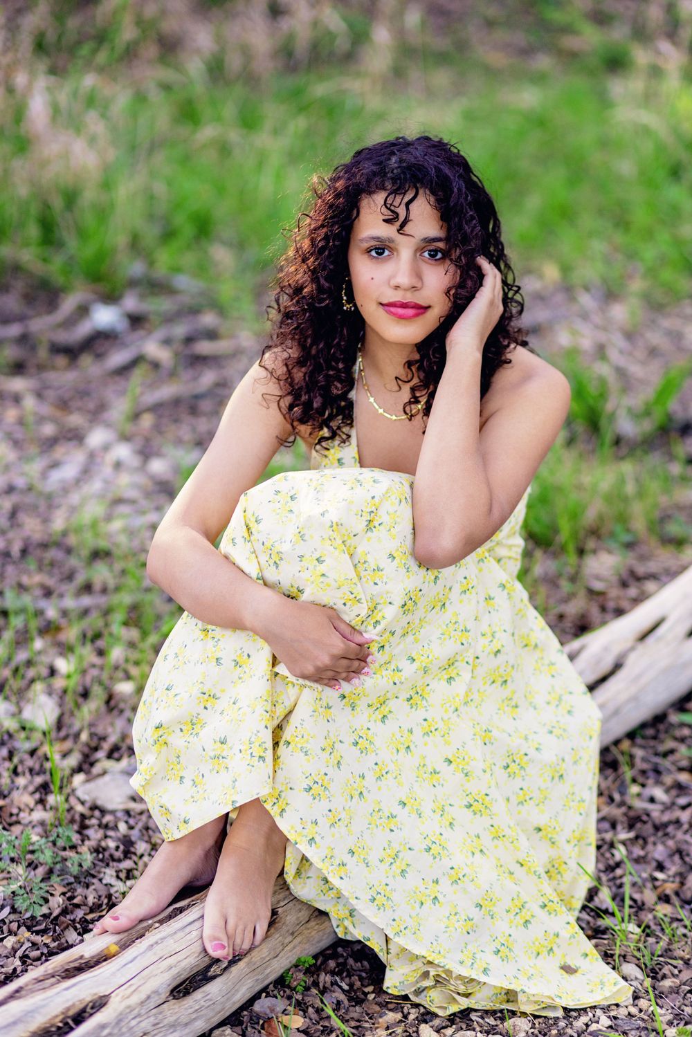 A person in a yellow floral dress sits on a fallen log outdoors with curly dark hair and natural scenery in the background.