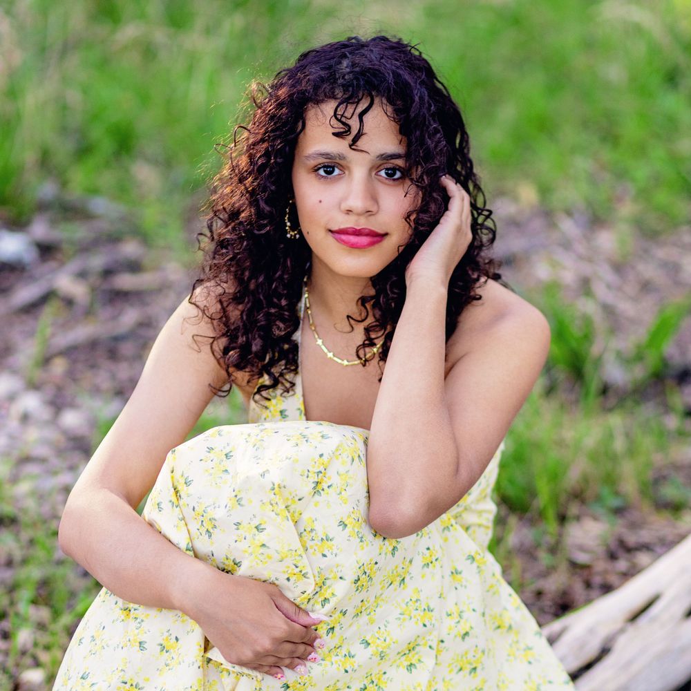 Person in yellow floral sundress poses outdoors among natural greenery with hand near face and curly hair.
