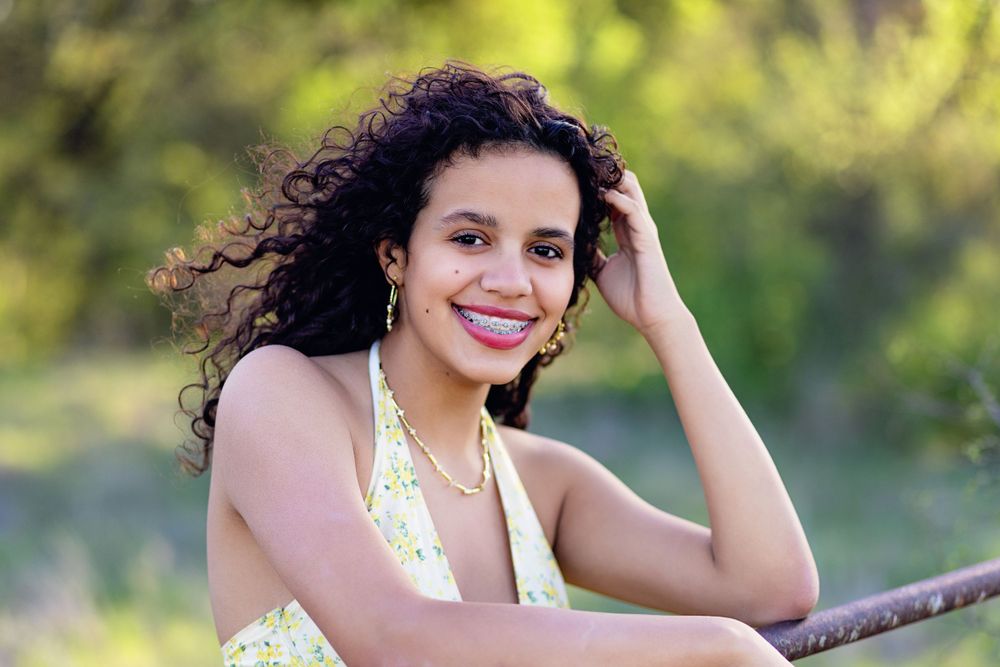Person with curly hair wearing a yellow dress sits outdoors in natural lighting with a warm smile.