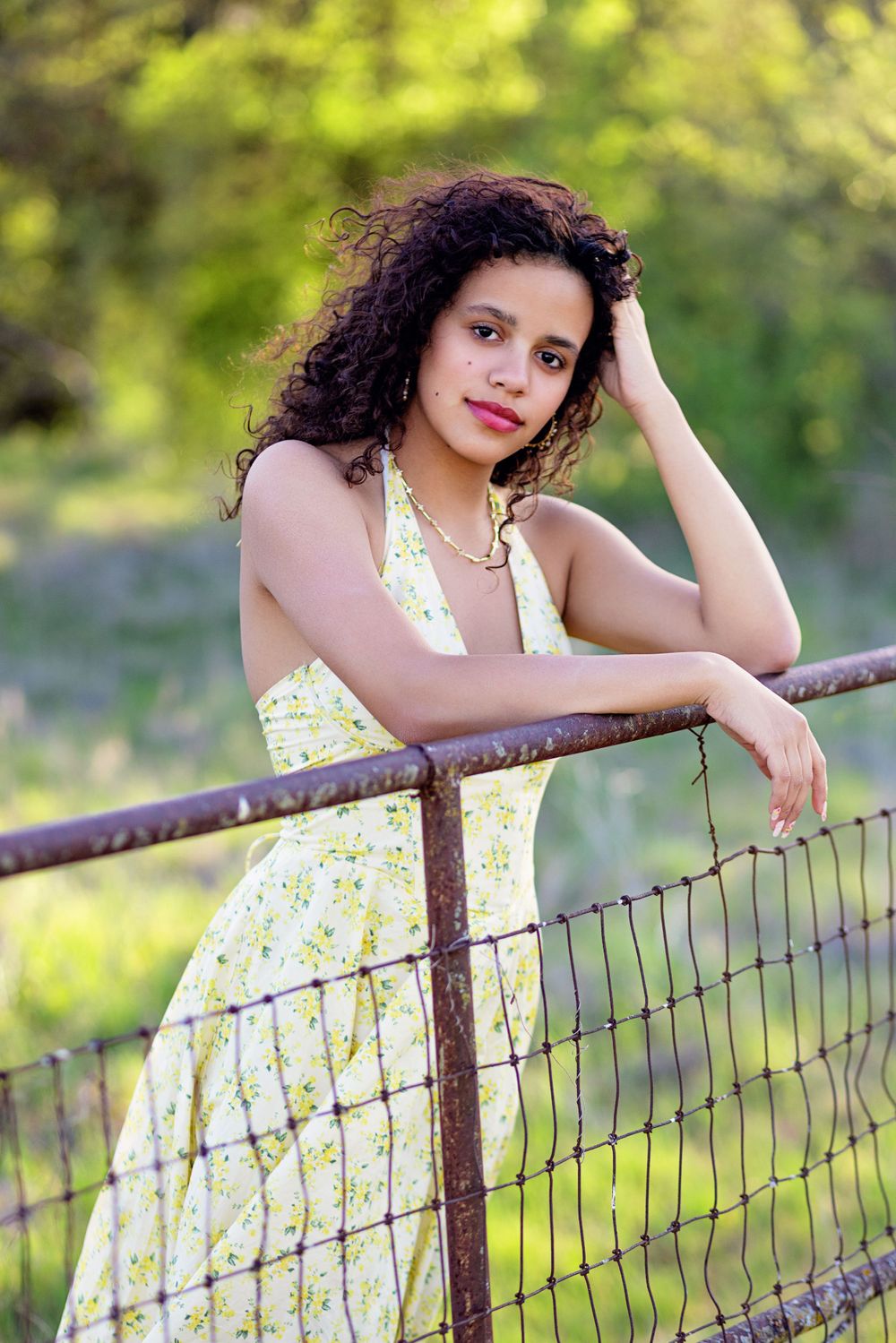 Woman in floral dress leans against rustic fence railing in outdoor summer portrait at golden hour.