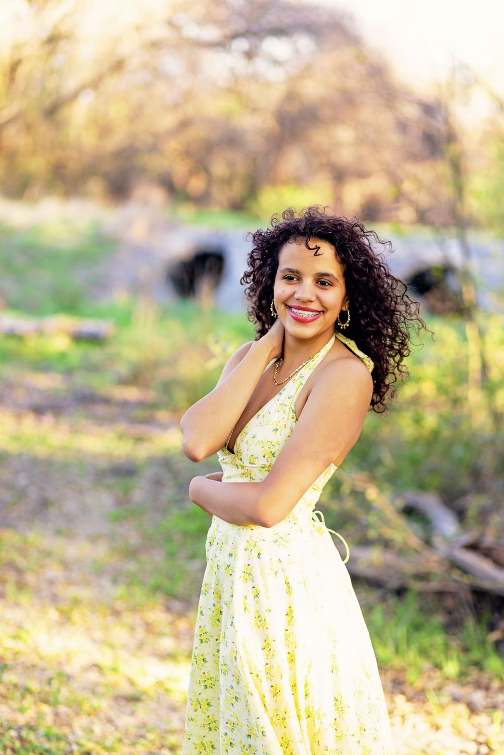 Someone in a white and yellow floral dress stands outdoors on a sunny day with natural curly hair and a warm smile.