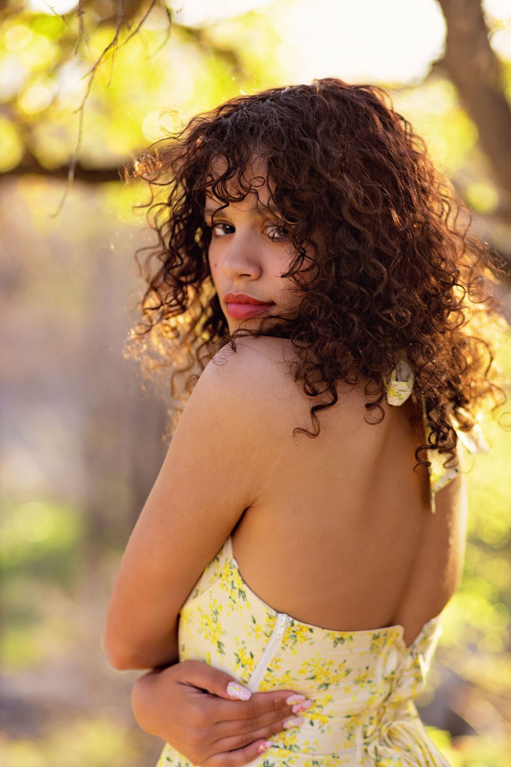 Portrait of a woman in a floral dress with curly dark hair looking over her shoulder in warm golden sunlight.