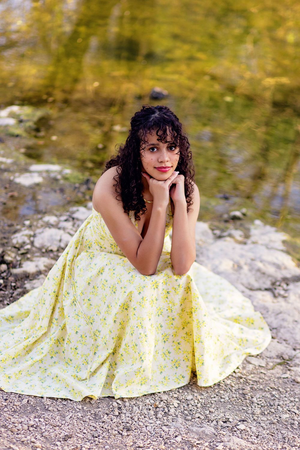 Person in a yellow floral sundress poses thoughtfully by a waterside on a rocky shore with autumn colors in background.