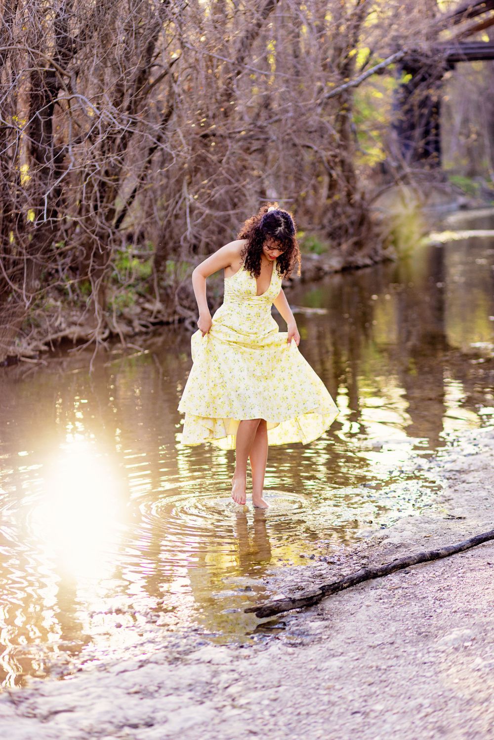 A person in a yellow polka dot dress stands at the edge of a tranquil stream, sunlight glinting off the water.