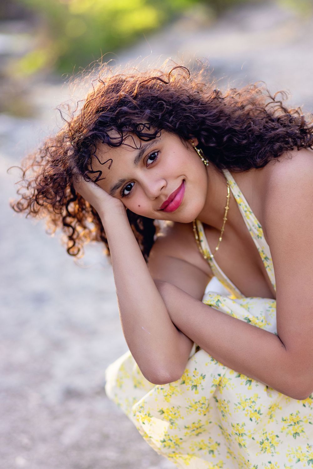 Portrait of someone in a yellow floral sundress with natural curly hair leaning on their hand against a blurred background.