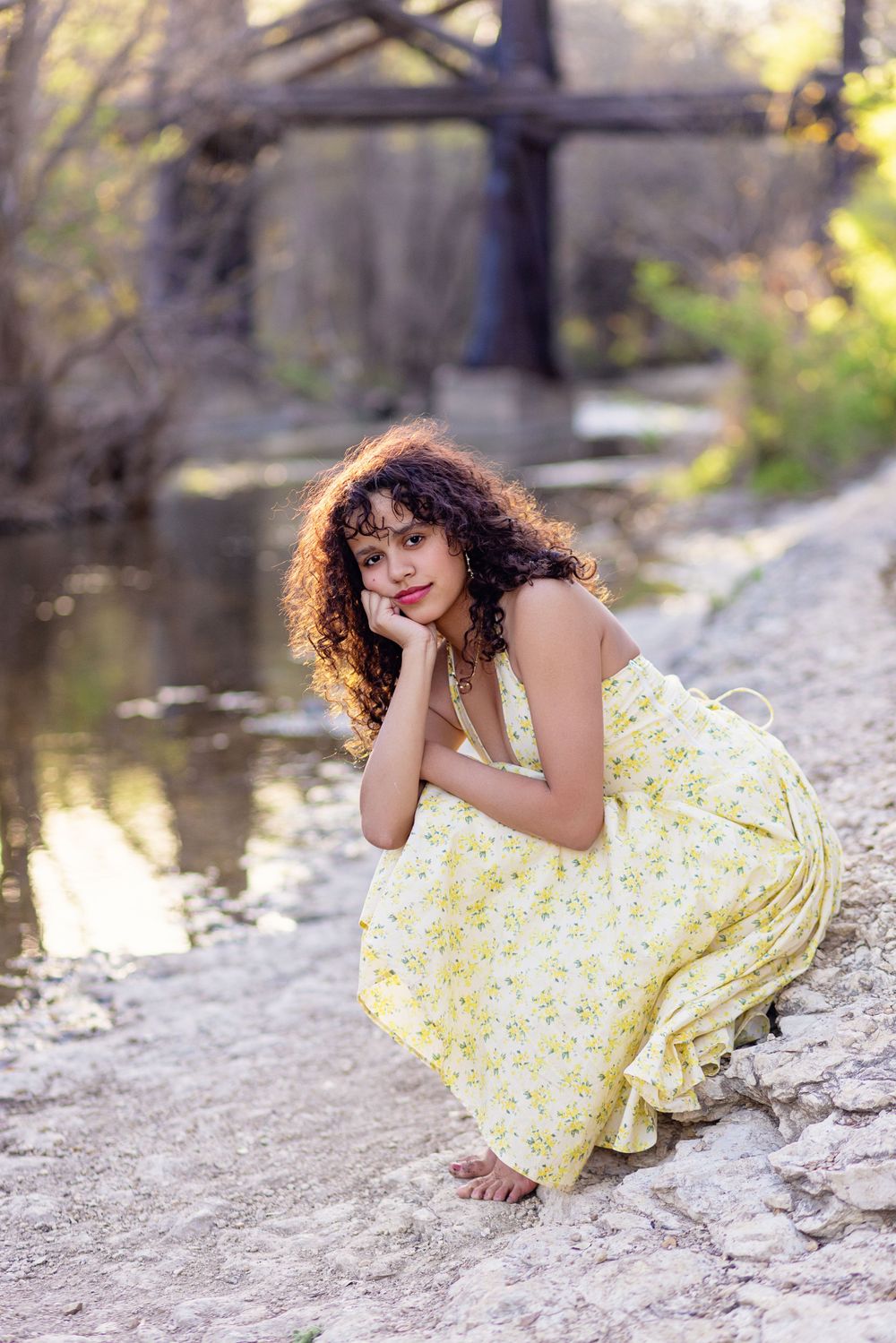 A yellow floral sundress flows gracefully by a creek with a bridge in the background during golden hour lighting.