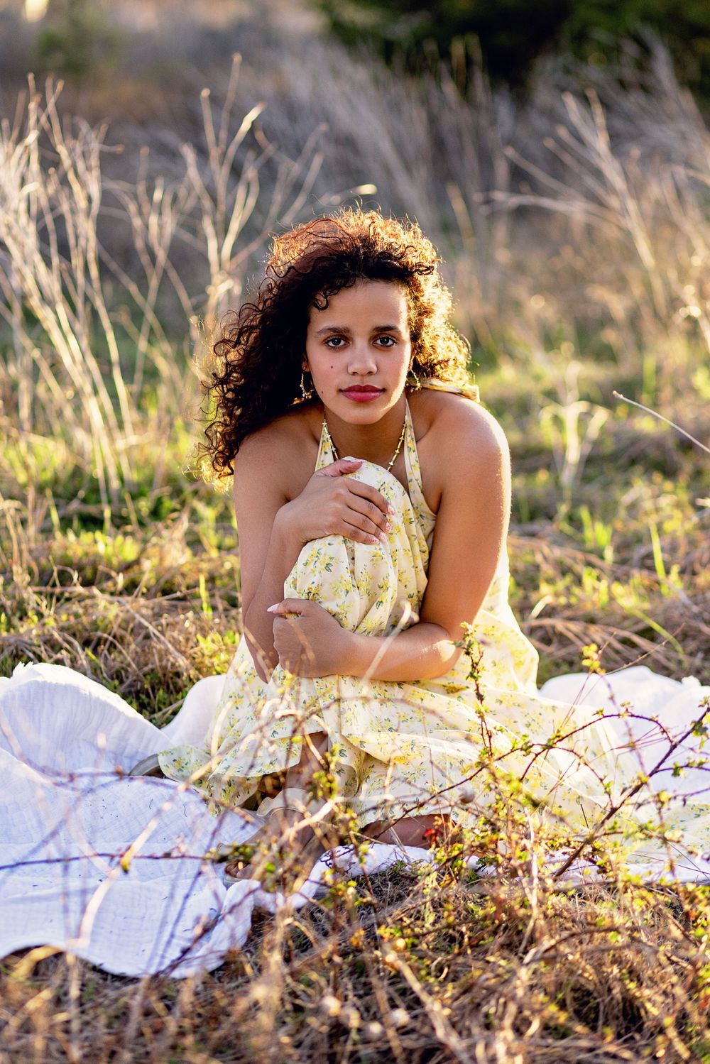 A person in a yellow floral dress sits gracefully on a white blanket amid sunlit tall grass during golden hour.