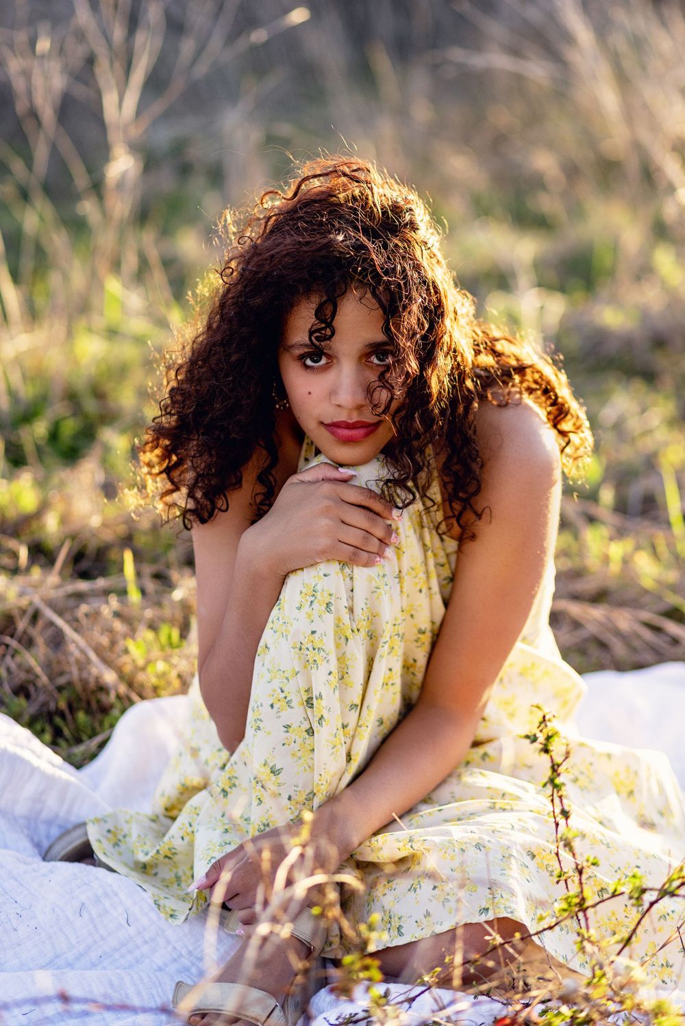Outdoor portrait with natural sunlight showing someone in a yellow dress sitting on a white blanket in tall grass.