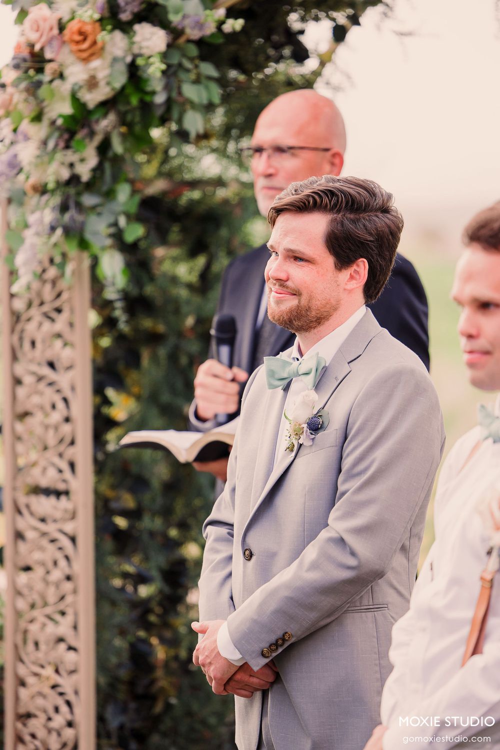 Wedding ceremony under floral arch with groomsmen in gray suits standing at altar.