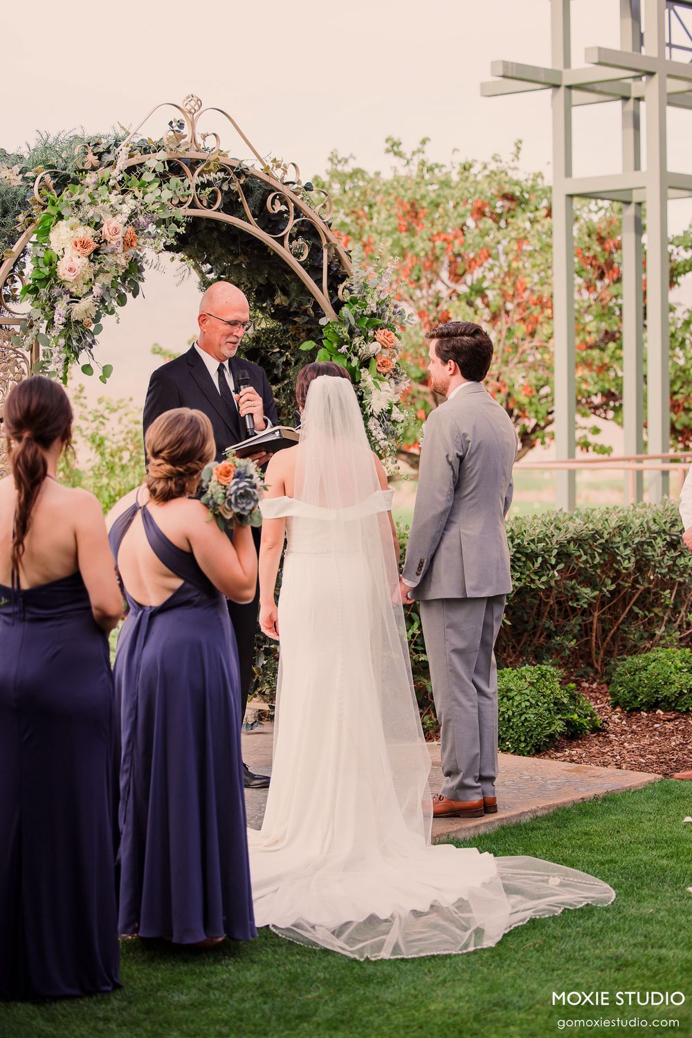 Wedding ceremony guests gather on a manicured lawn with flowering bushes and white pergola.