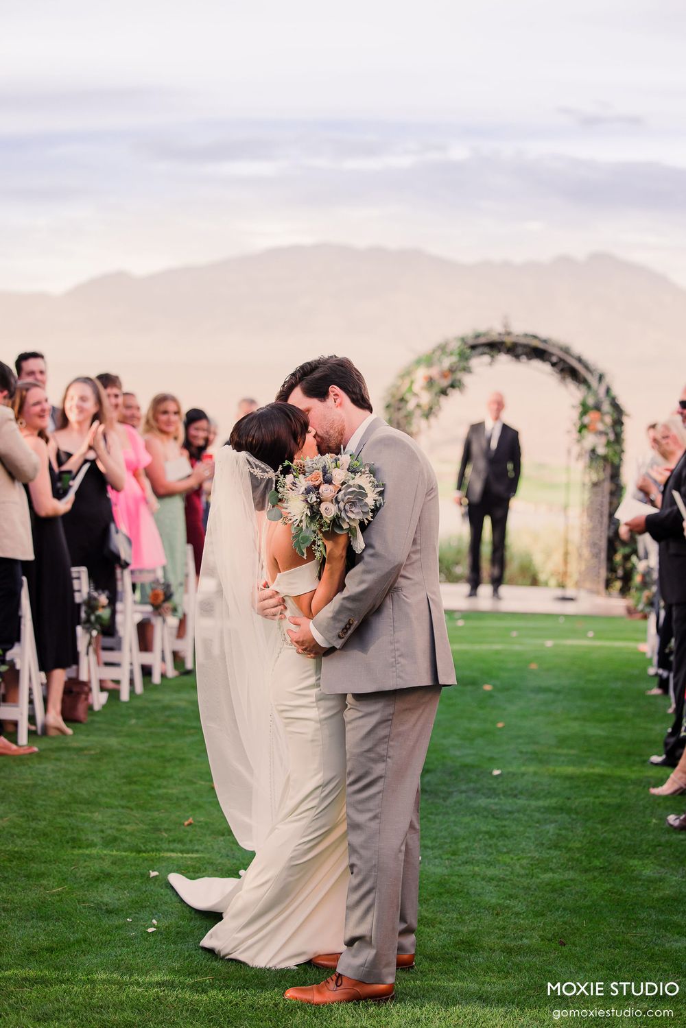 A romantic moment between newlyweds kissing under a floral arch as guests look on at an outdoor ceremony.