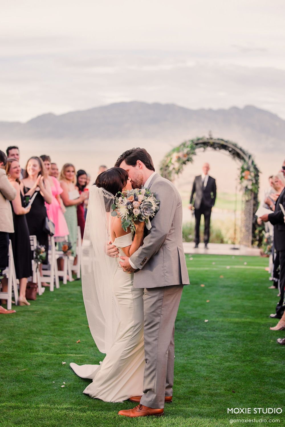 Romantic outdoor wedding ceremony on a green lawn with mountain backdrop and floral arch decor.