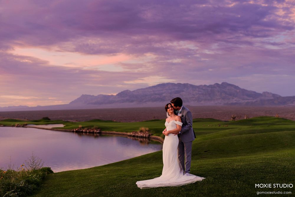 A wedding couple embraces on a golf course with purple mountains and dramatic sunset sky in the background.