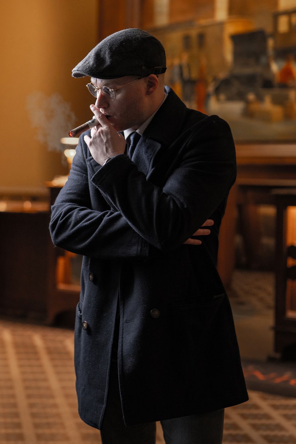 Man in black coat and flat cap smoking a cigar indoors, looking thoughtful in warm ambient lighting.