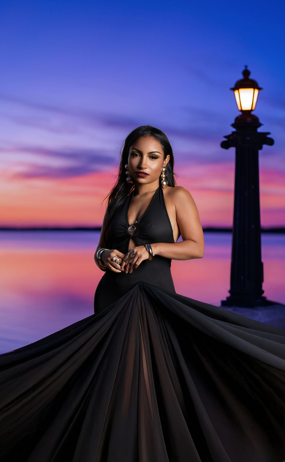 A silhouetted lighthouse stands against a vibrant purple and orange sunset sky as a figure in a flowing black gown poses nearby.