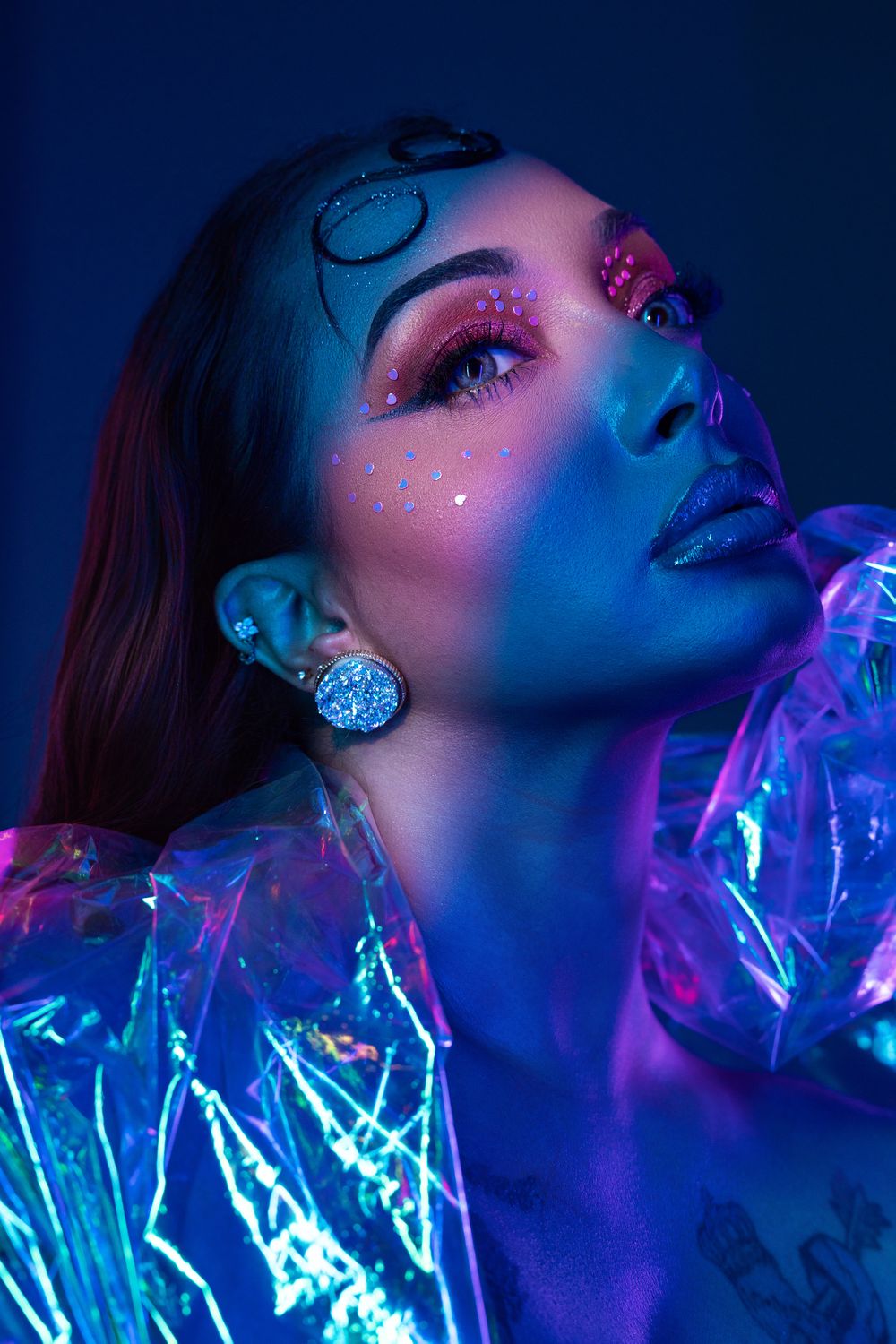 Woman with neon UV makeup, holographic jacket, and glitter gems under blue and pink studio lights.