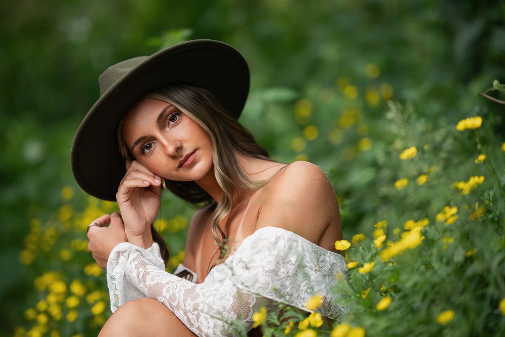 Young woman in wide-brim hat and white off-shoulder lace top sitting among yellow wildflowers in lush green garden.