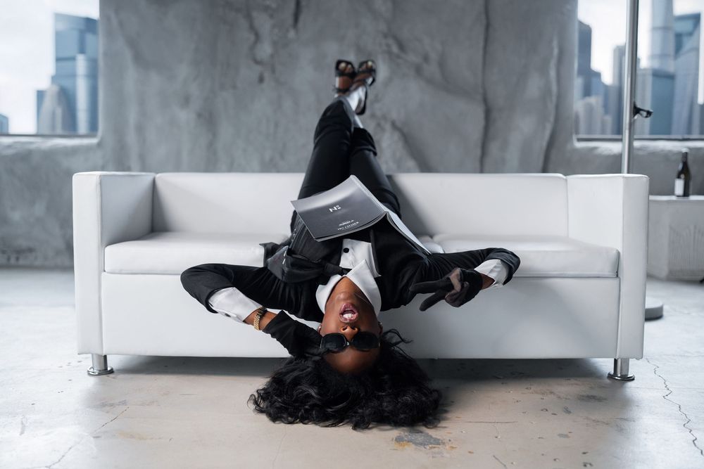 A dancer in black attire performs an artistic pose while lying upside down on a white couch in an industrial space.