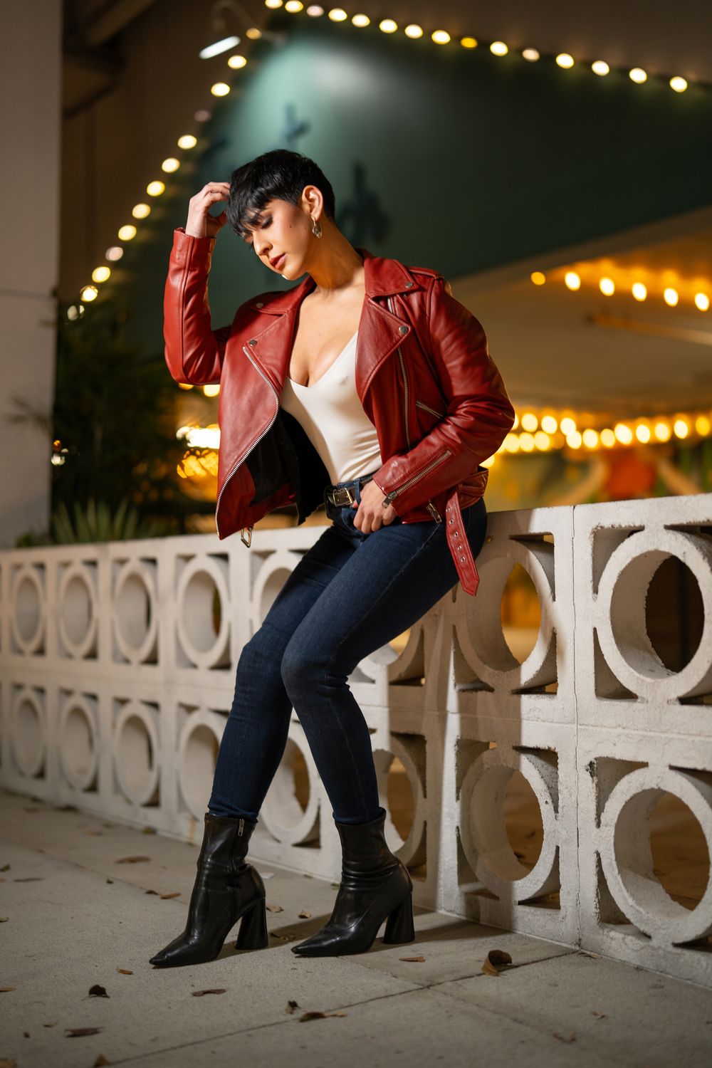 A person in a red leather jacket and jeans poses on a decorative white wall at night with string lights in the background.