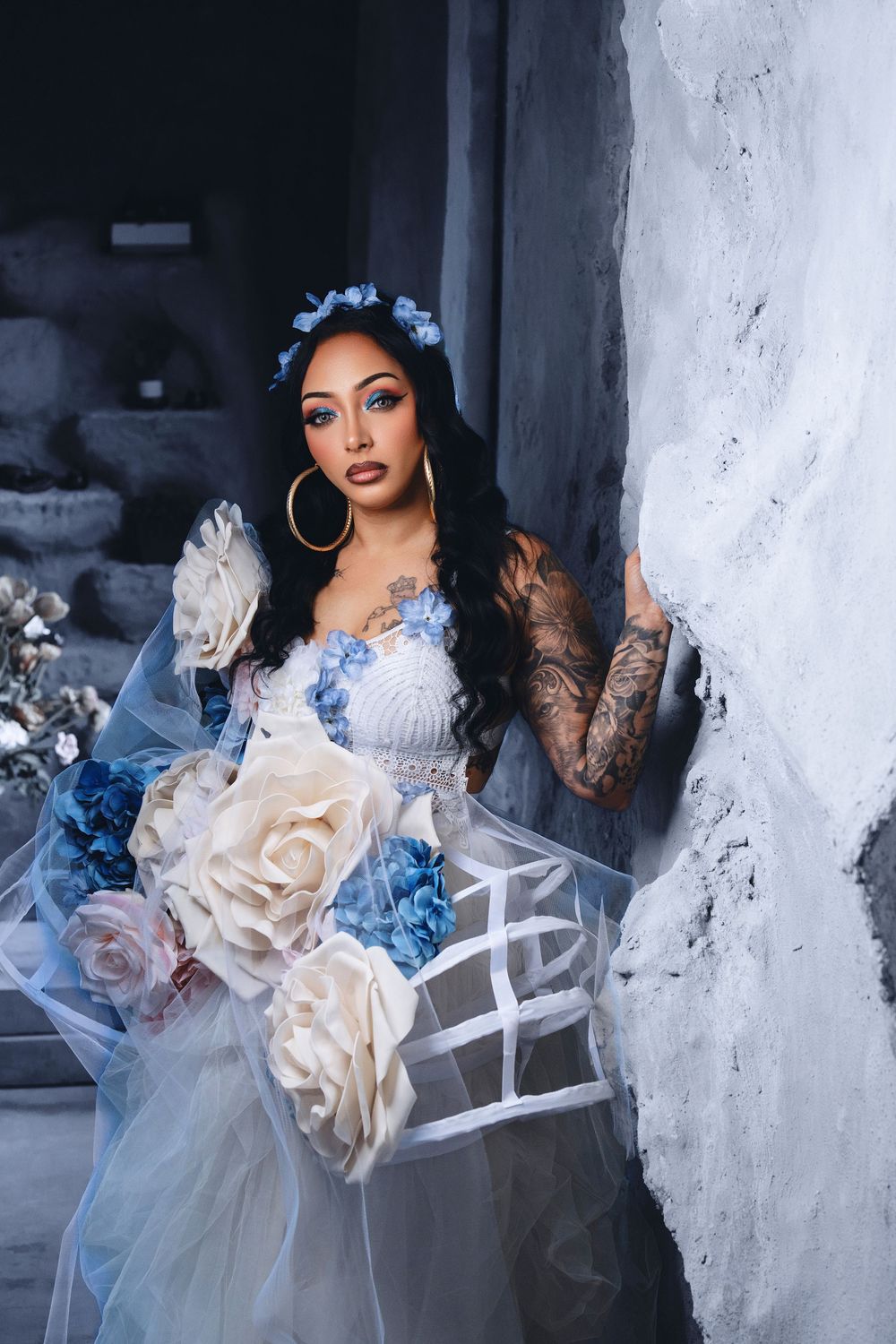 Woman in blue floral gown with tattoos and hoop earrings holds large roses near textured gray wall.