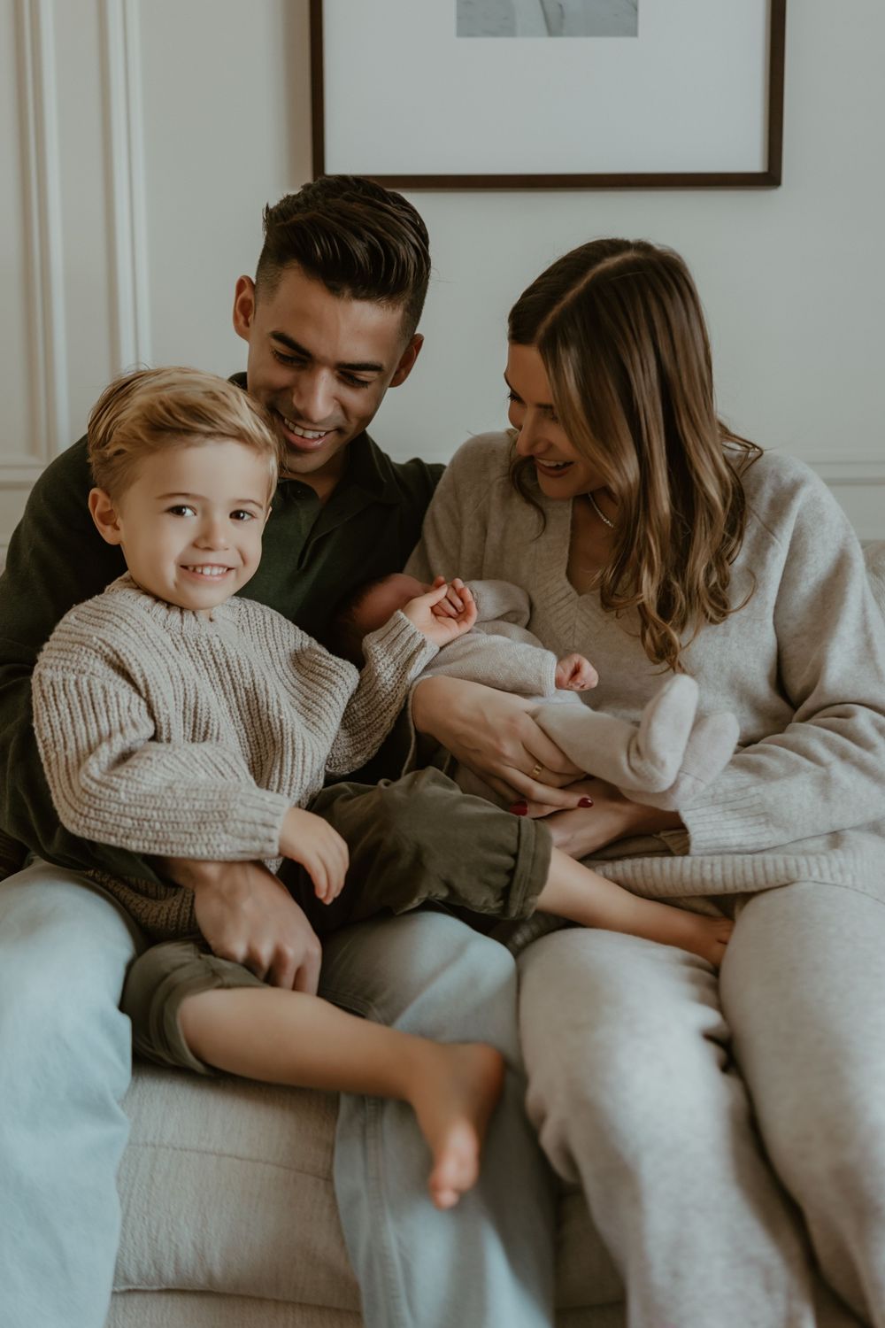 Family lounging together on white couch wearing cozy neutral sweaters and casual attire.