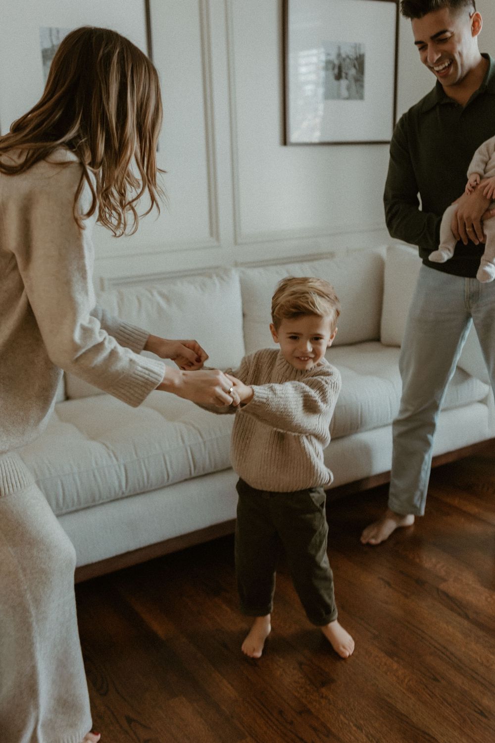Family in neutral sweaters sharing playful moments together on hardwood floors by a white couch.