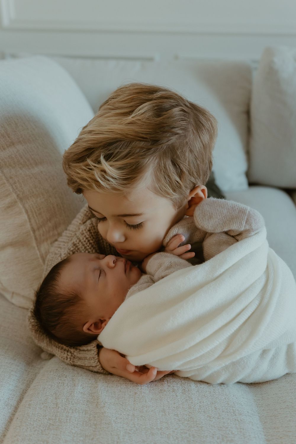 Two siblings share a tender moment on a couch as the older child gently embraces the younger one.