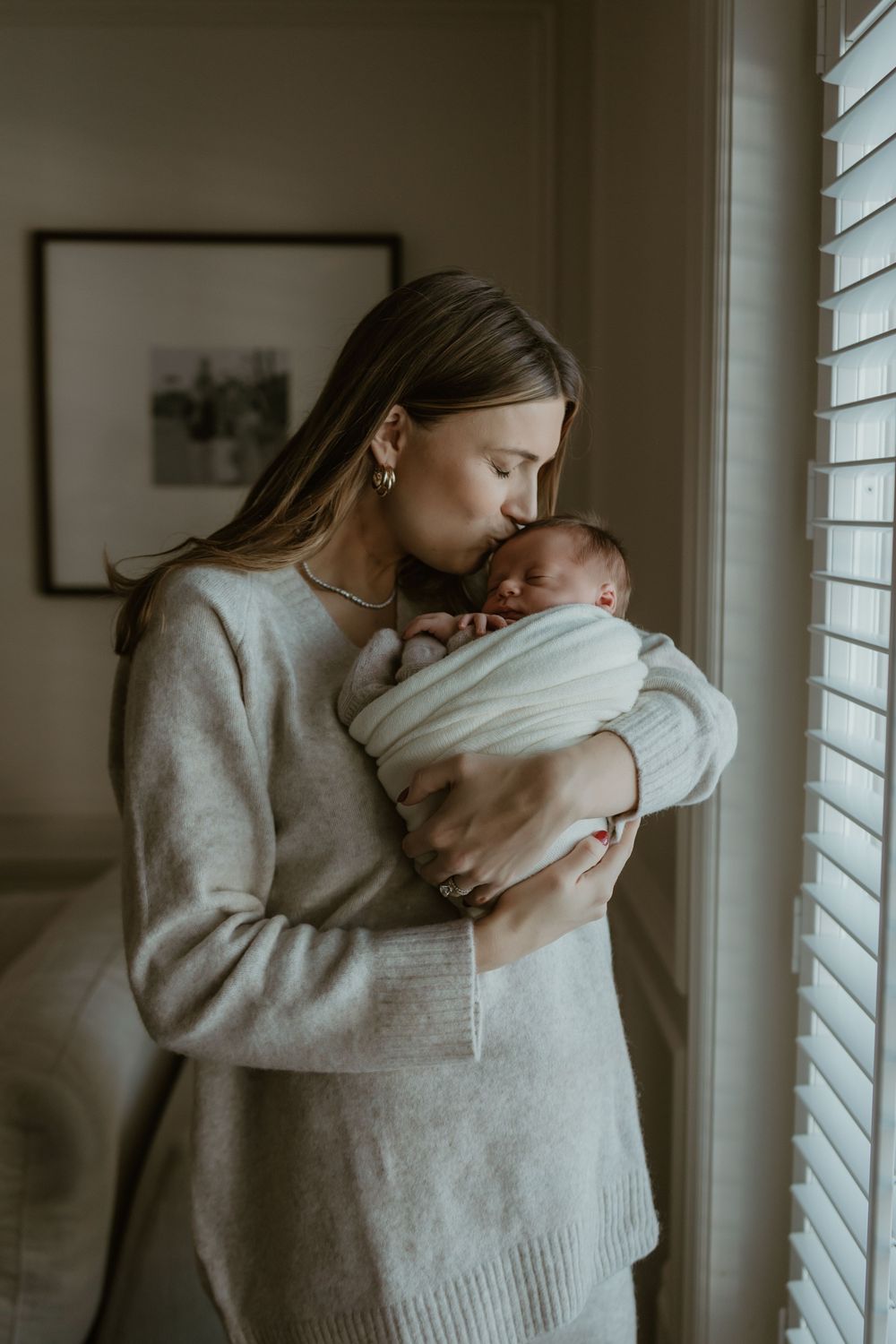A tender moment by a window as a parent in a cozy sweater embraces their sleeping newborn in soft natural light.