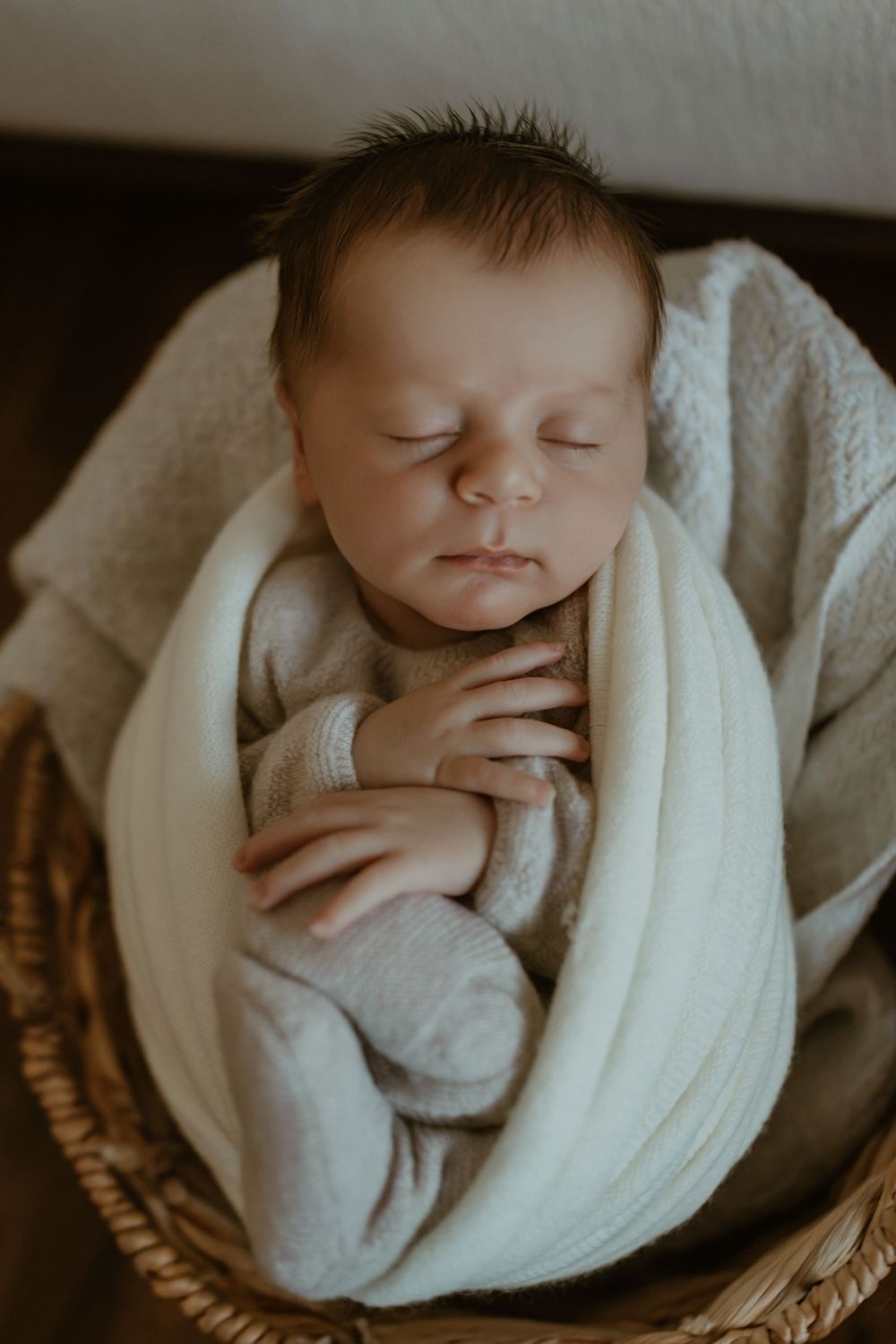 A peaceful sleeping newborn swaddled in a soft white blanket rests in a woven basket.