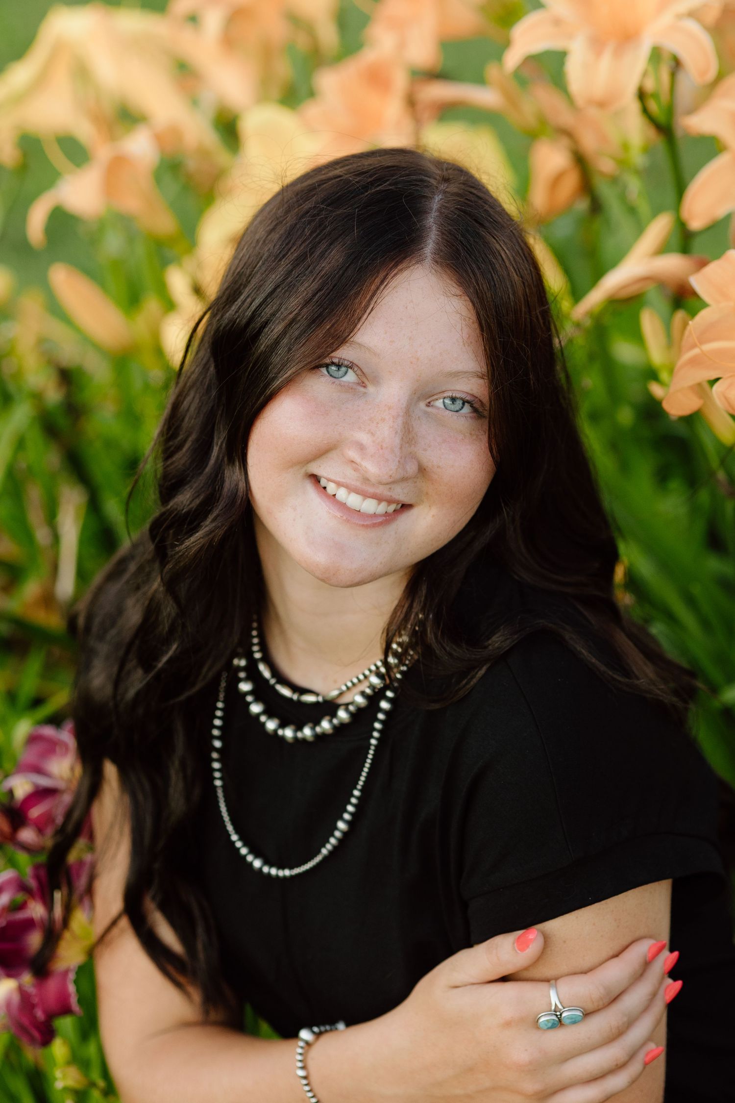 Smiling dark-haired woman with blue eyes poses in front of vibrant orange lilies.