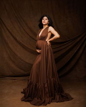 Maternity portrait against brown backdrop featuring a flowing chocolate brown gown with ruffle details and empire waist design.