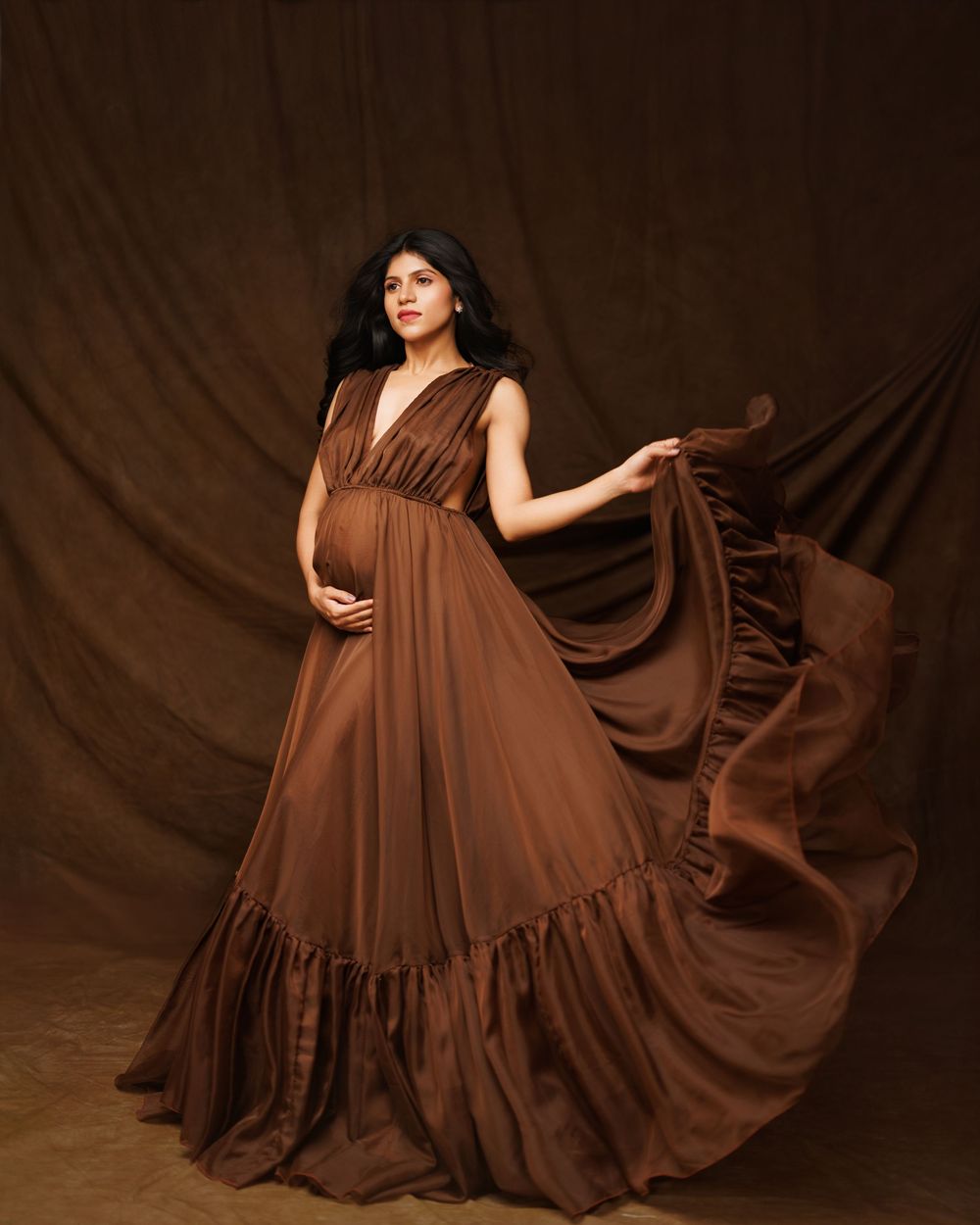 A flowing brown maternity gown with empire waist billows dramatically against a dark studio backdrop.