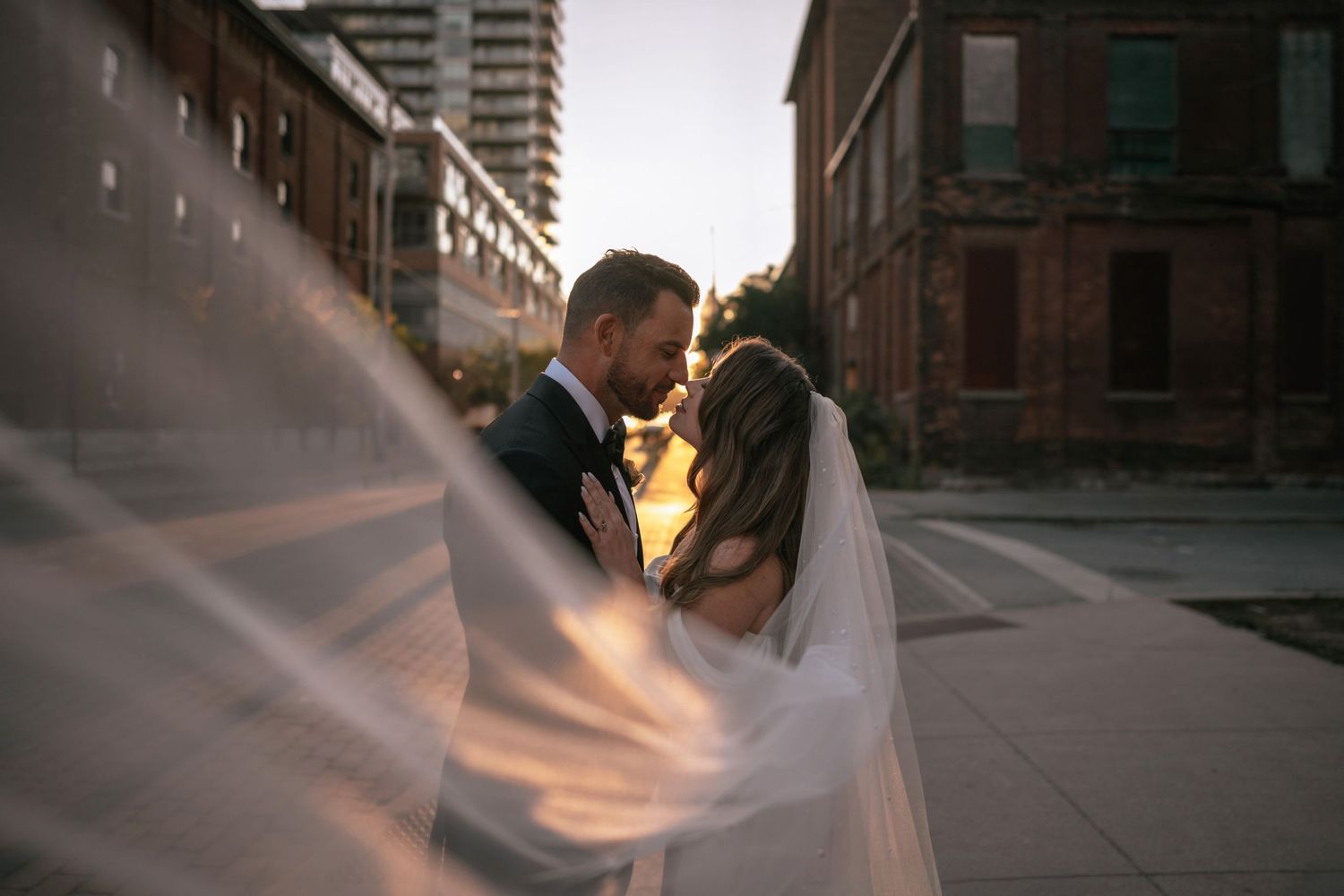 A romantic wedding couple embraces in golden sunlight on an urban street as the bride's veil flows in the wind.