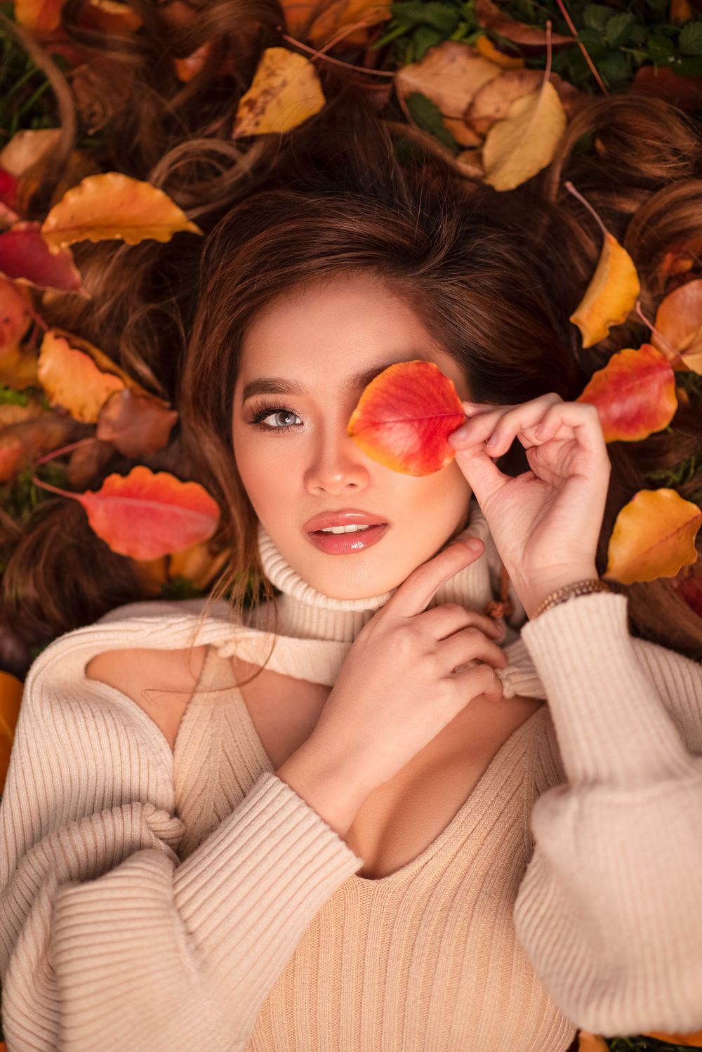 Woman in a cream sweater posing among colorful autumn leaves with a red leaf held over one eye.