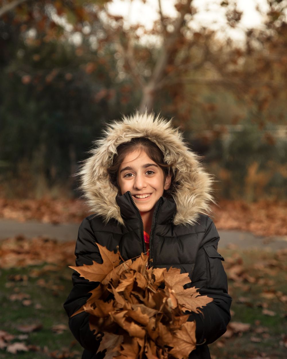 Smiling child in a fur-trimmed winter coat holding a bundle of autumn leaves outdoors.
