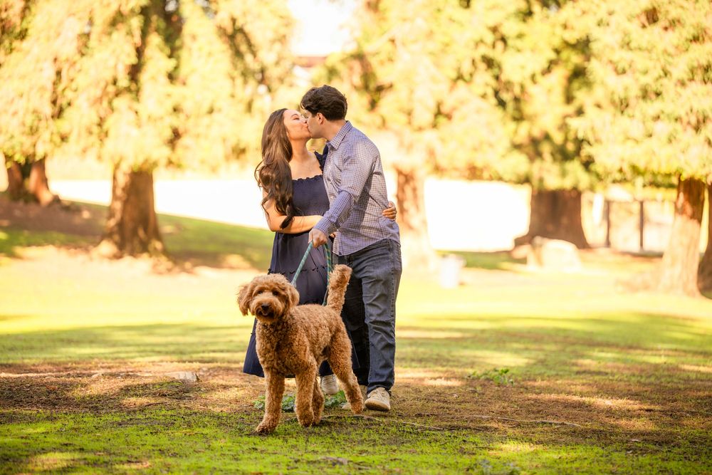 Couple kissing in a sunlit park with their dog during a fall photography session.