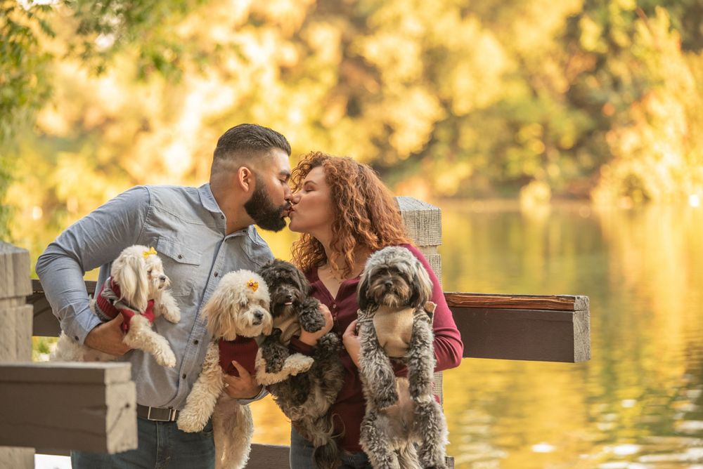 Couple sharing a kiss beside a lake while holding their dogs during an outdoor portrait session.
