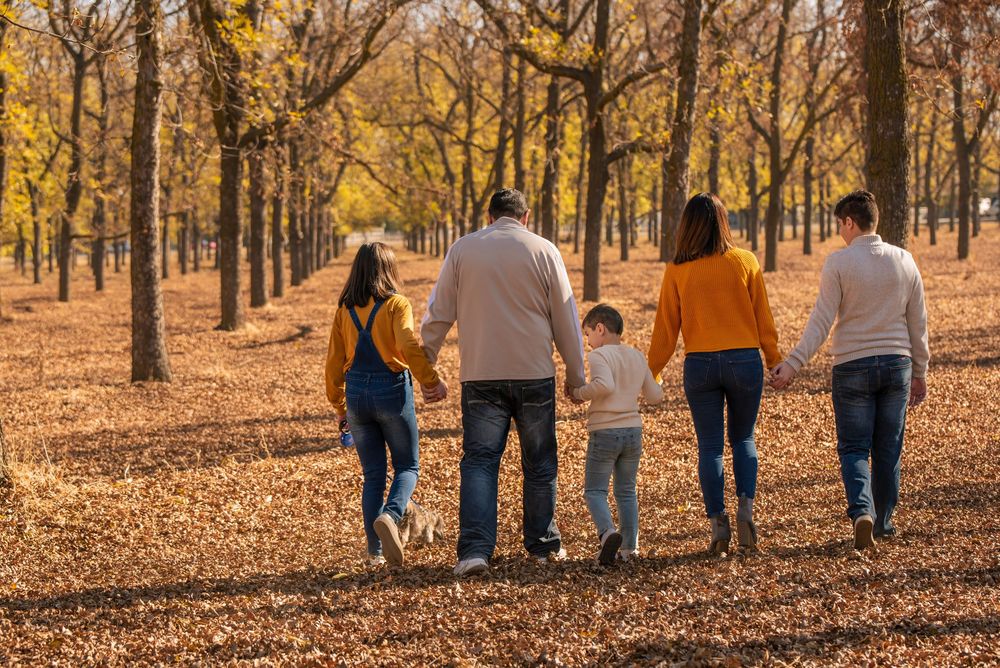 Family of five walking hand in hand through a tree-lined path covered in autumn leaves.