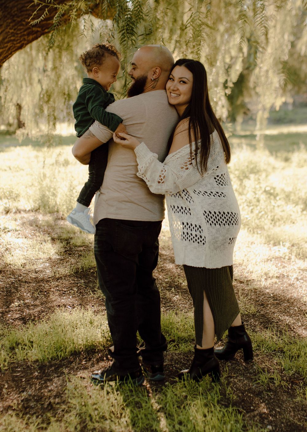 Family shares tender moment under willow tree in warm sunlight.