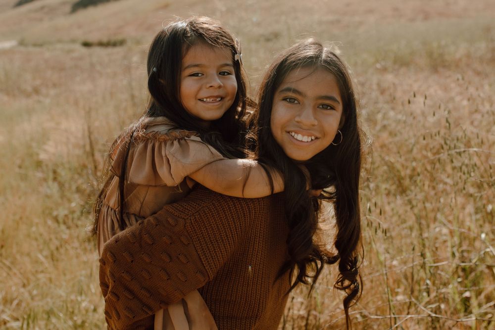 Golden sunset photo of two girls in brown sweaters posing in grassy field.