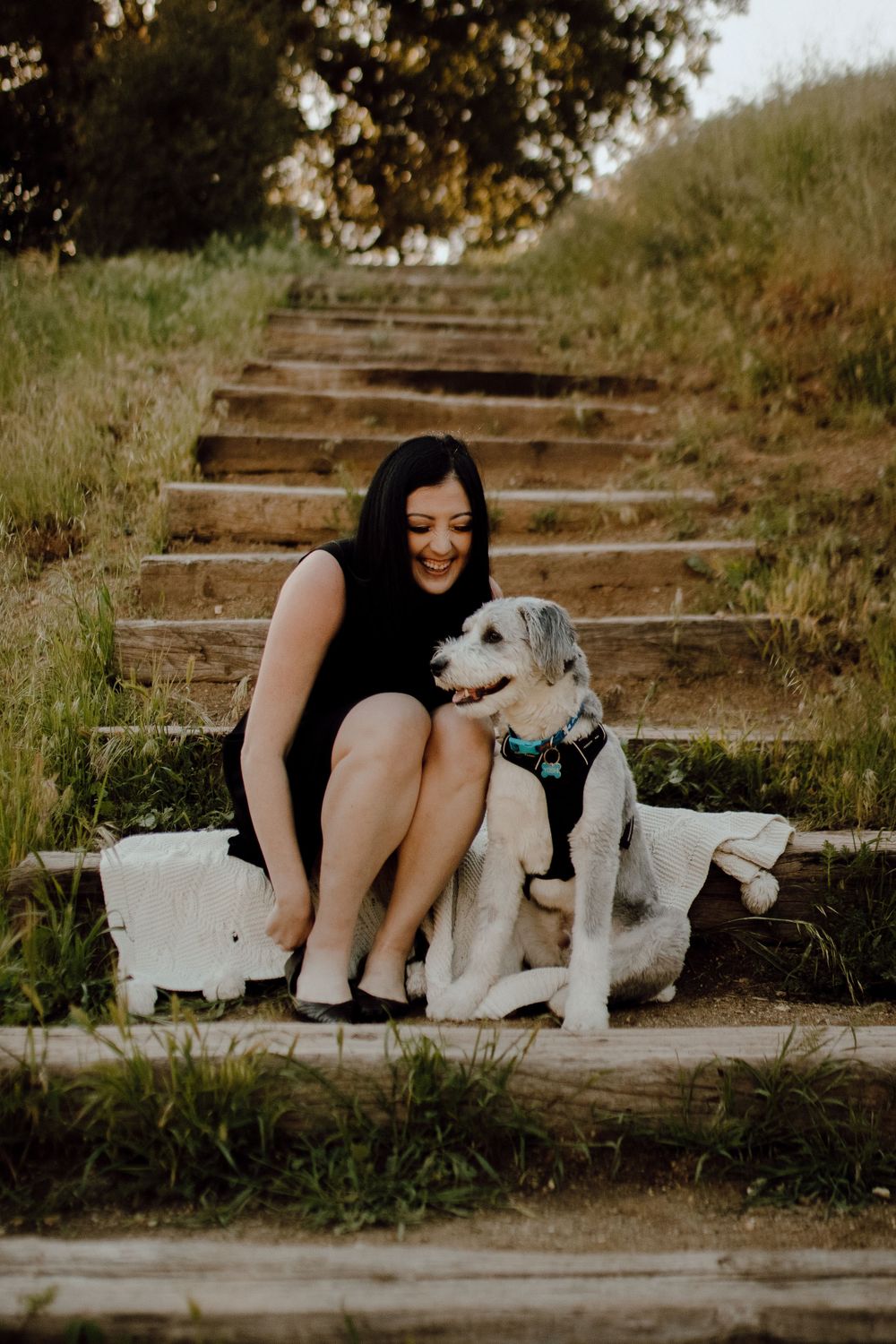 Person in black dress sits with happy white dog on outdoor stone steps.