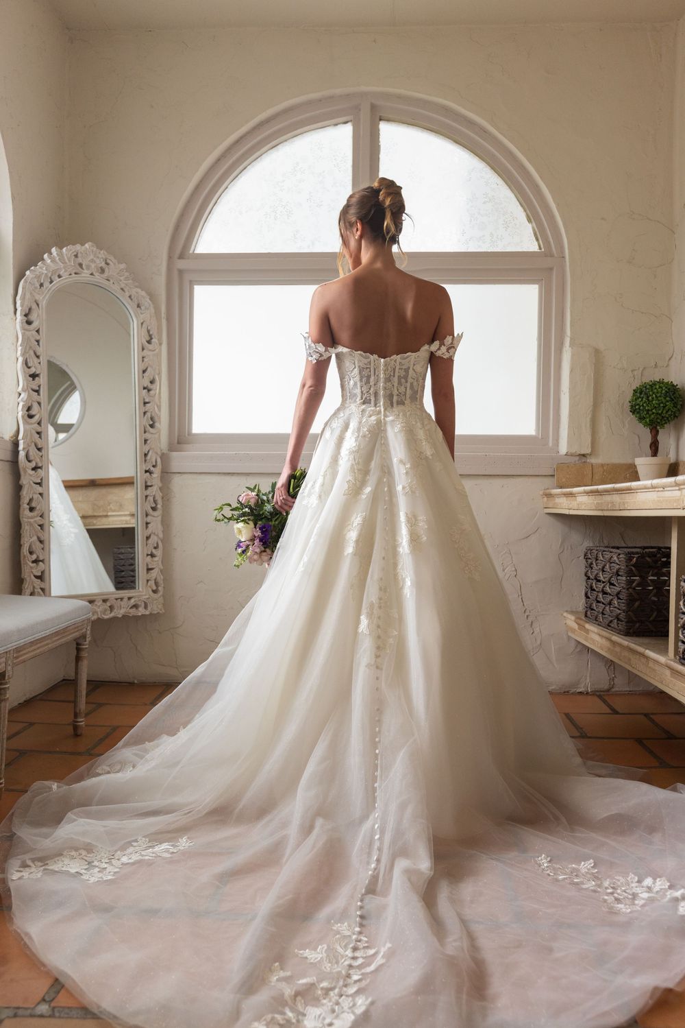 Elegant off-shoulder wedding gown with lace details and dramatic cathedral train shown from behind in sunlit bridal room.
