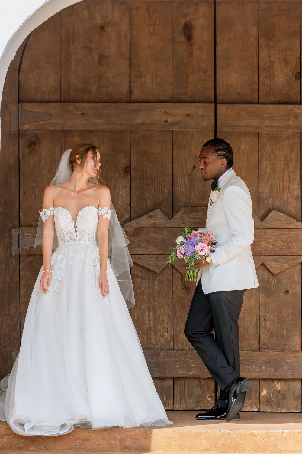 A bride in a white off-shoulder gown and groom in white tuxedo jacket share a playful moment against rustic wooden doors.