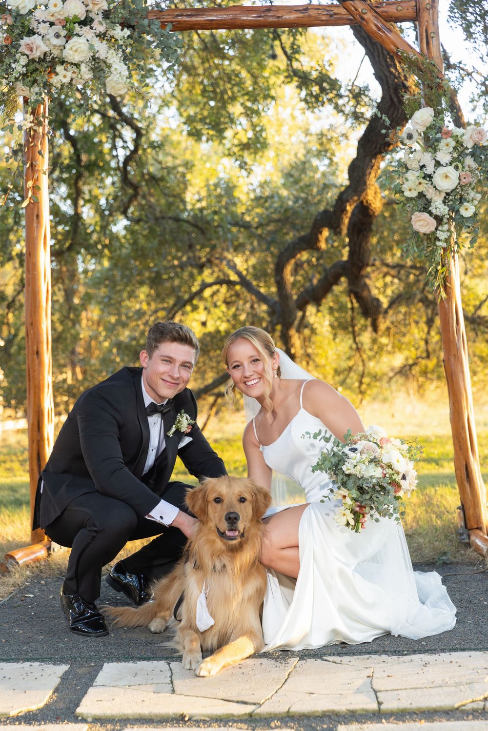 A couple in wedding attire poses with their golden retriever dog under a rustic wooden pergola at sunset.