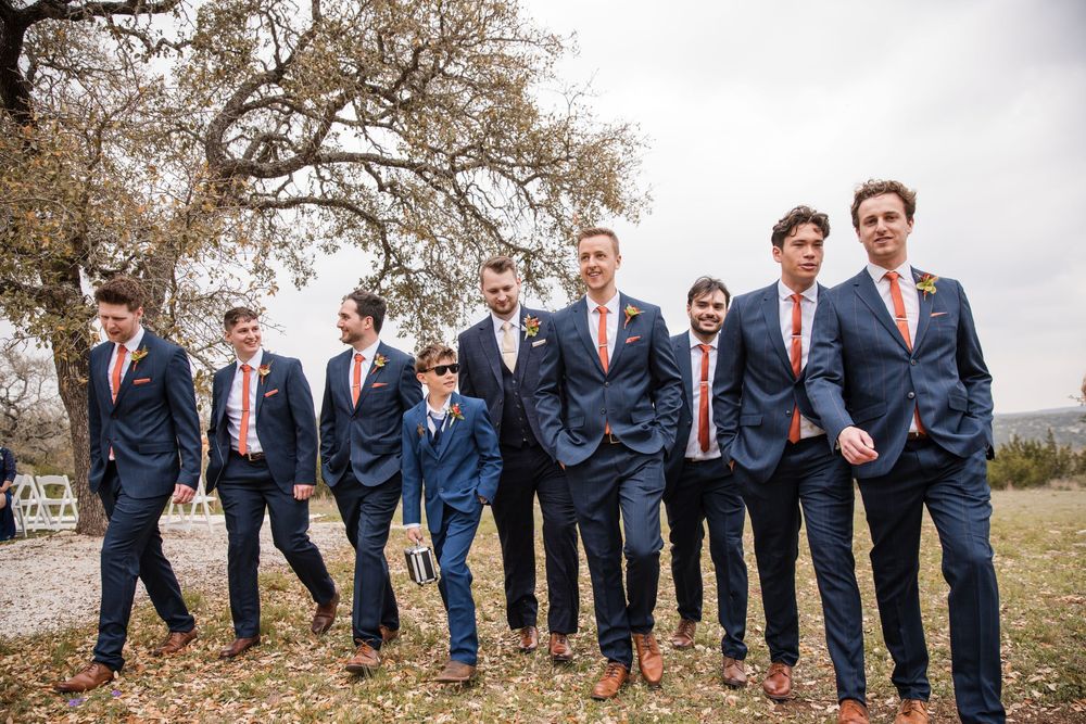 Groomsmen in navy suits pose playfully in a row outdoors for wedding photos on a cloudy day.