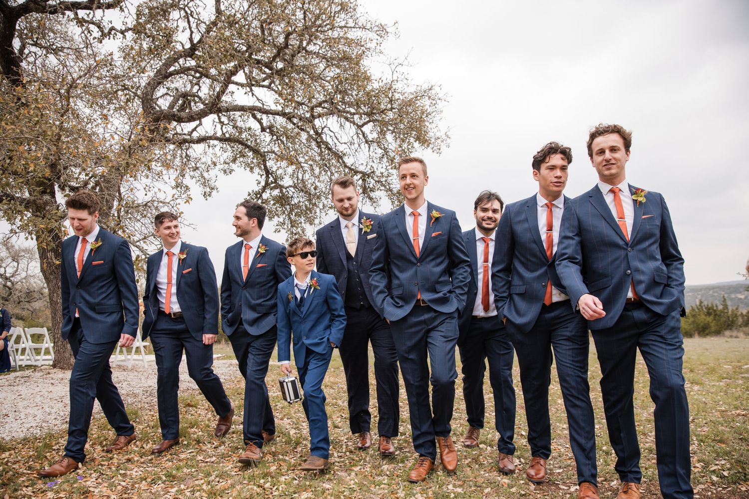 Groomsmen in navy suits pose playfully in a row outdoors for wedding photos on a cloudy day.