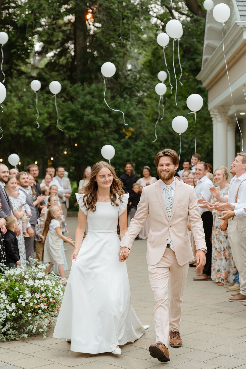 Wedding guests line a walkway decorated with white balloons as a couple walks through during their celebration exit.