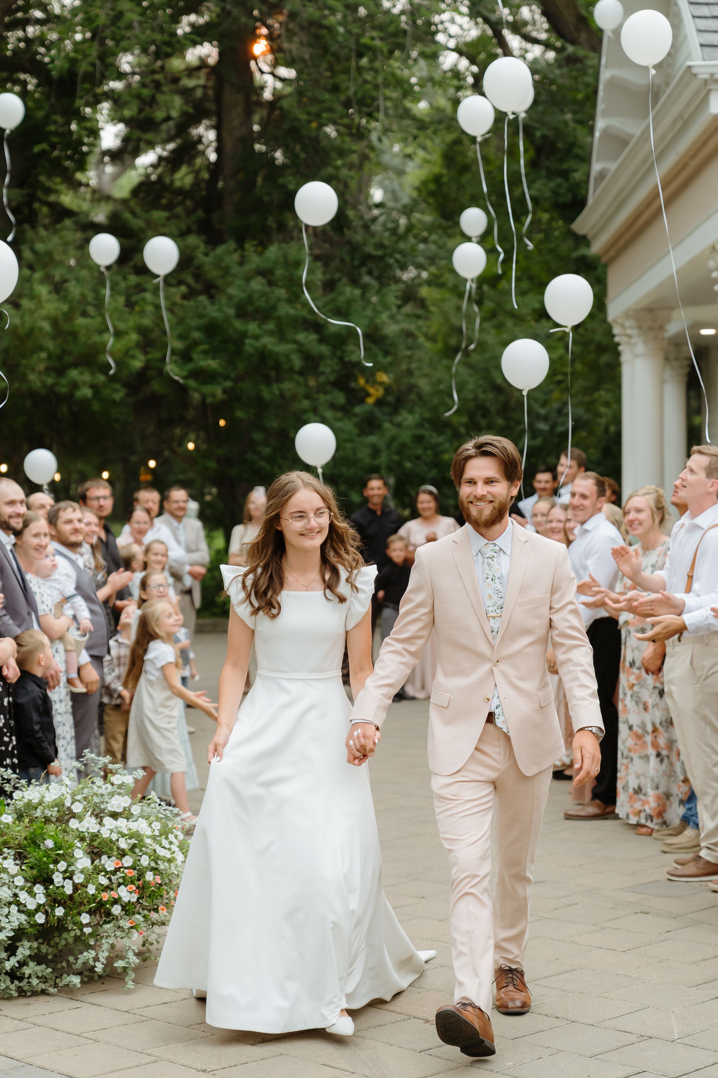 Wedding guests line a walkway decorated with white balloons as a couple walks through during their celebration exit.