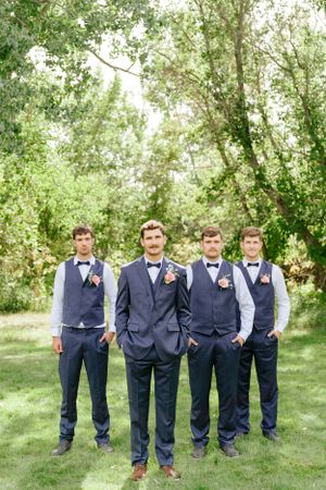 Four groomsmen in navy suits and vests pose for a formal photo under green trees.