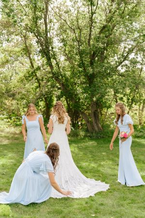 Wedding party arranging the bride's lace train on grass under trees.